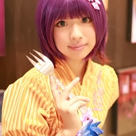 A female cosplayer with a purple wig in her booth selling photos of herself modeling.