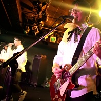 Guitar player ヒット君 (Hit-kun) of 装置メガネ (Sōchi Megane) singing with his band mates.