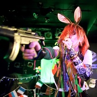 Singer ハル (Hull) of マイナス人生オーケストラ (Minus Jinsei Orchestra) pointing a toy gun at the audience.