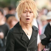 Japanese guy with blond hair and a black coat looking quite surprised.