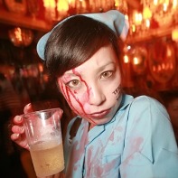 Japanese girl in a light-blue nurse outfit covered with fake blood holding a beer.
